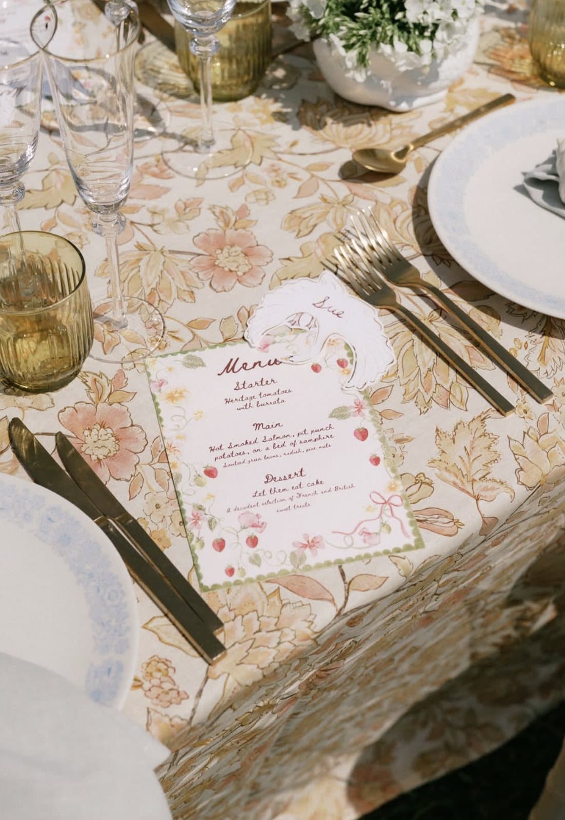 A hand-painted menu card on a floral cloth, with gold cutlery and amber glassware